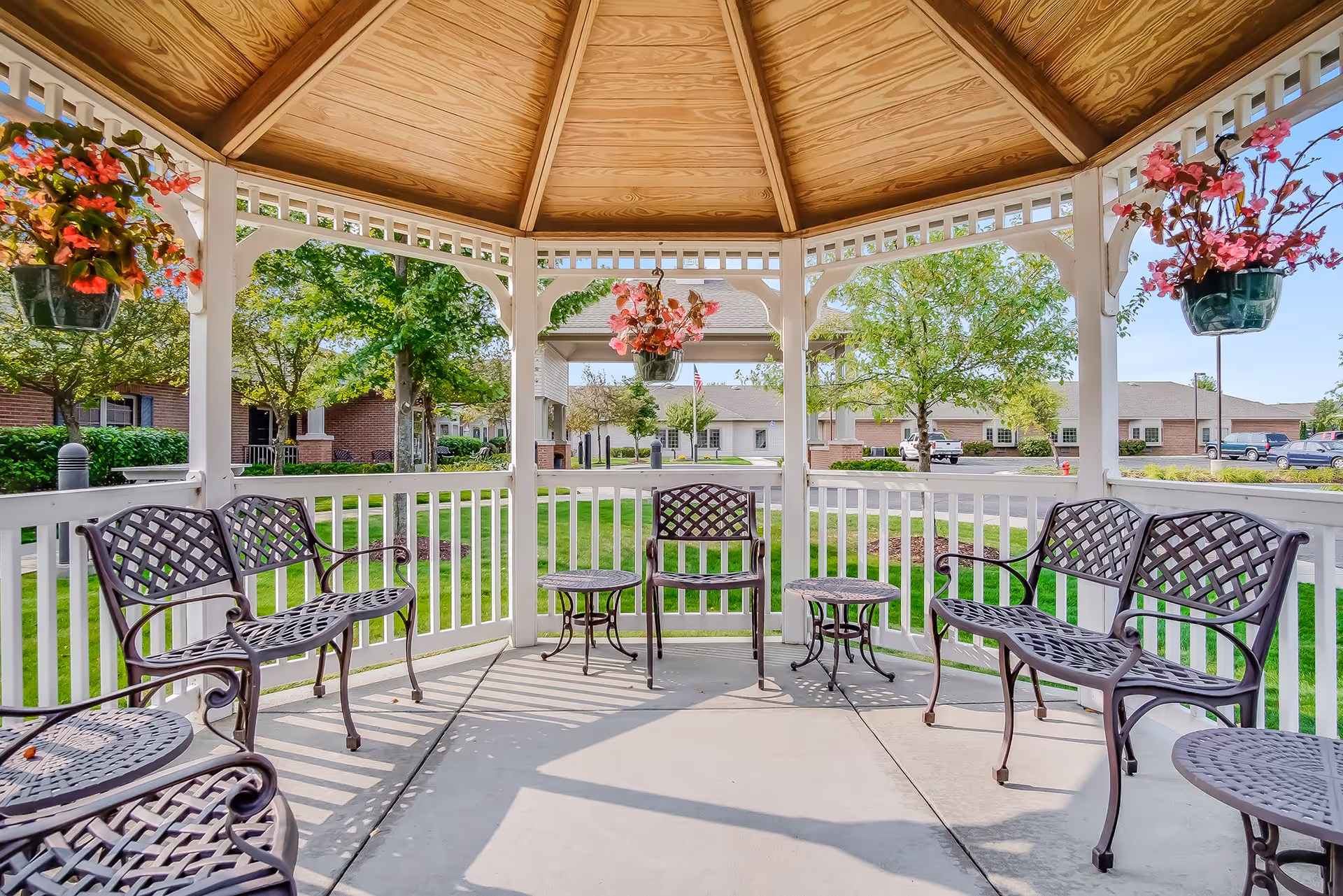 View from inside a wooden gazebo with a peaked roof, featuring metal benches and small tables arranged around the perimeter. Hanging flower baskets with pink and red flowers decorate the gazebo. Outside, there is a well-maintained lawn, trees, and buildings in the background under a clear blue sky.