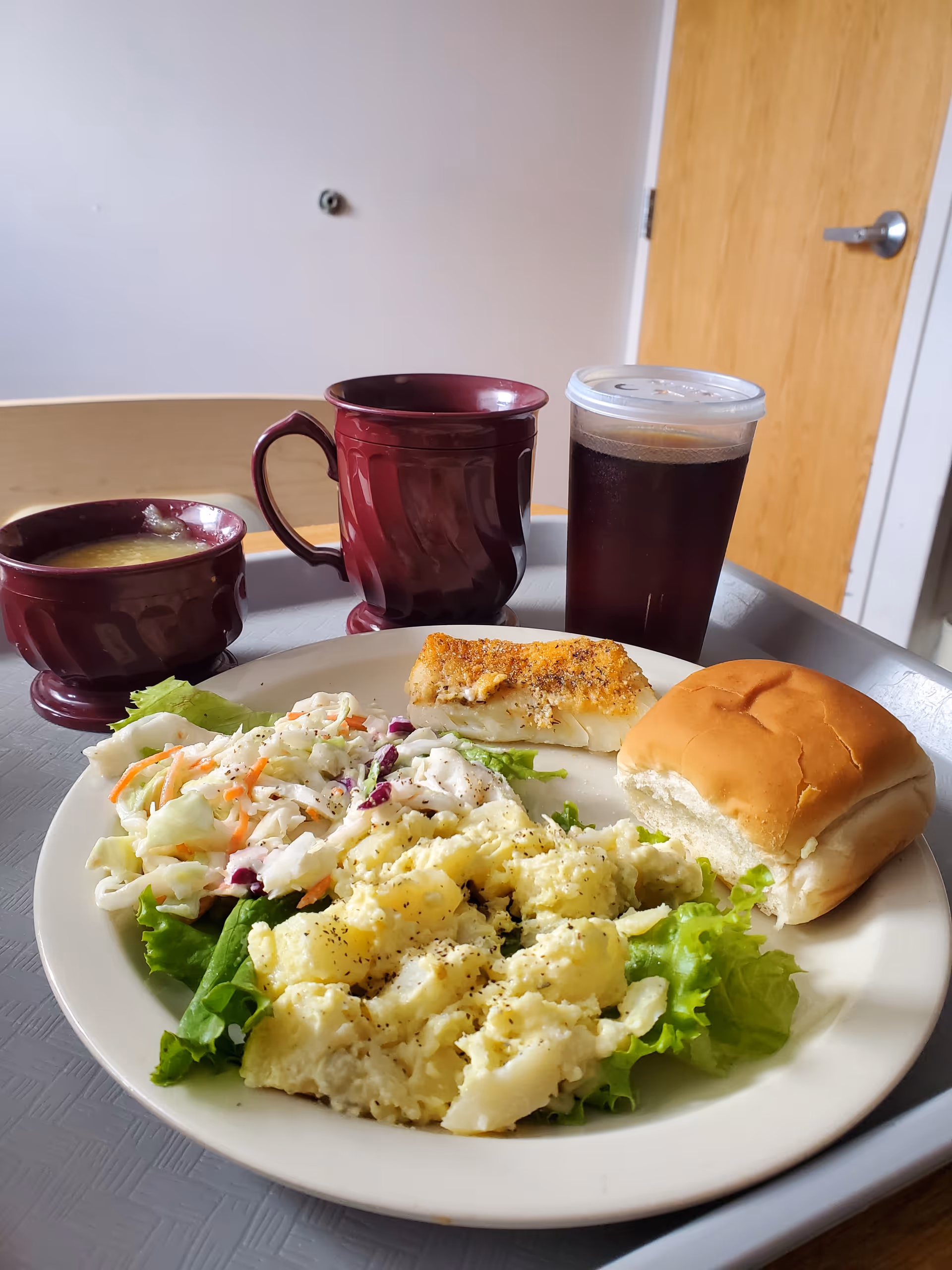 Tray with mashed potatoes, coleslaw, fish, and a bun on a dining room table.