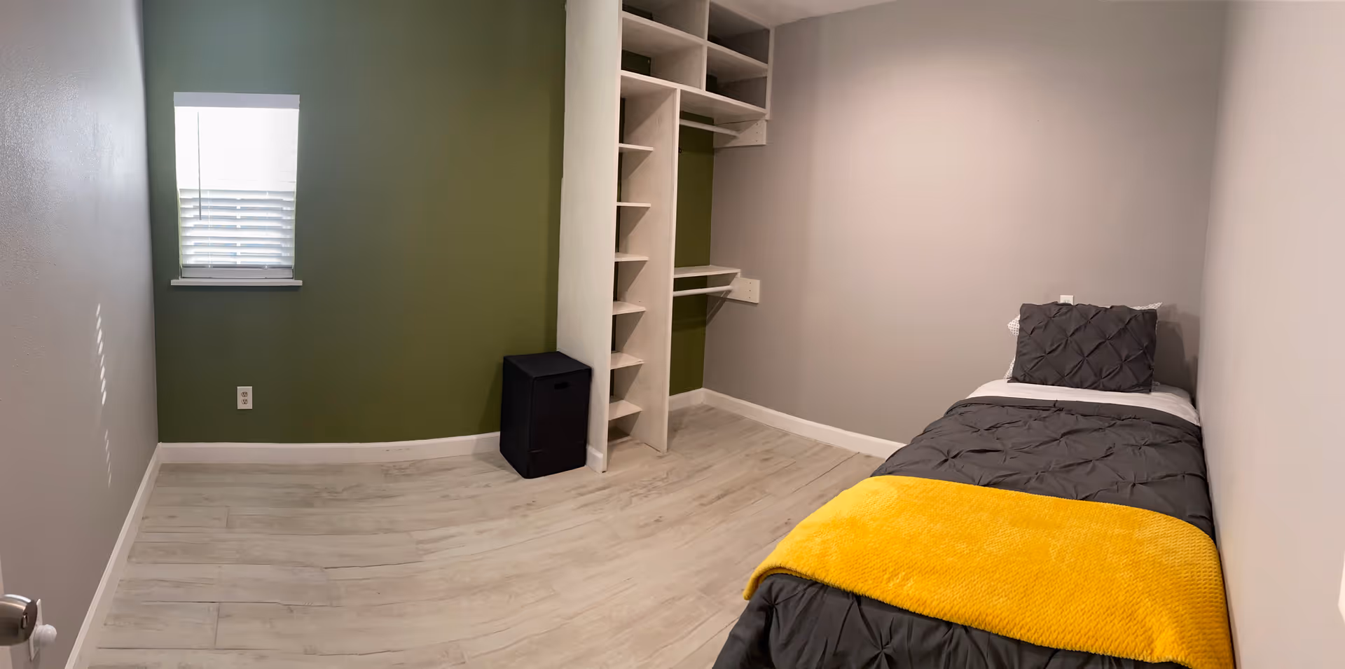 A small bedroom with a bed, closet shelves, window, and light wood flooring.