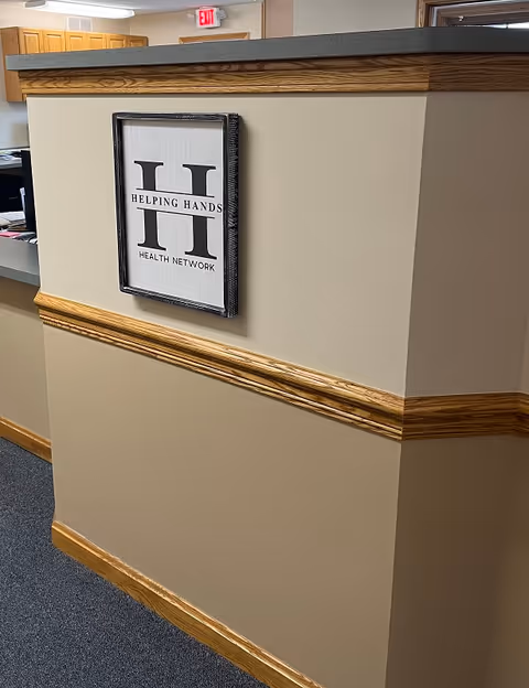 Interior hallway/reception area with beige walls, wood trim, and a Helping Hands Health Network sign.