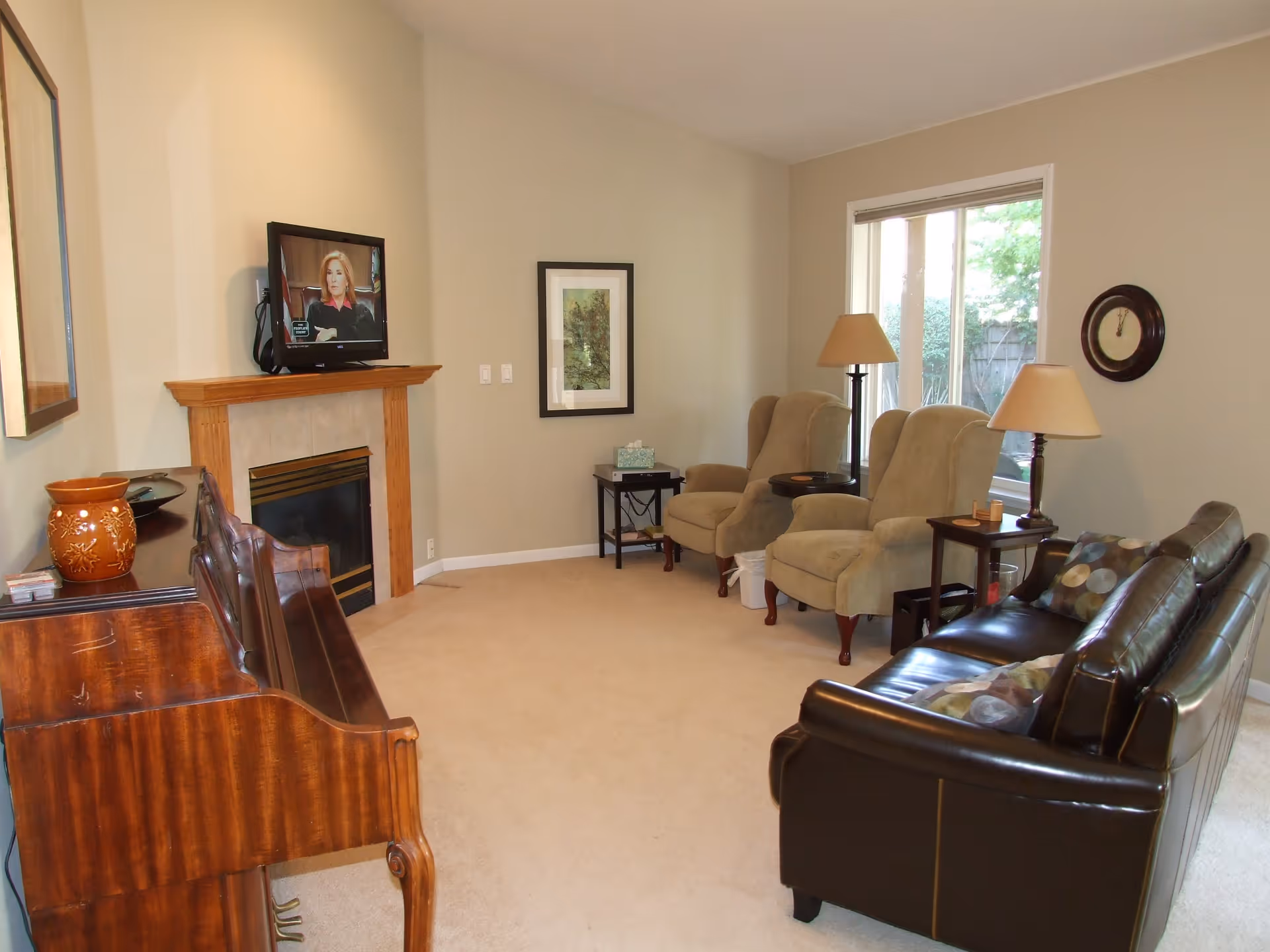 A cozy living room with beige walls and carpet. The room features a wooden piano on the left, a fireplace with a mounted TV above it, two beige armchairs with side tables and lamps, a dark brown leather sofa with patterned cushions, a wall clock, and a window showing greenery outside.