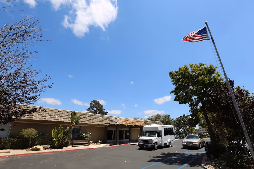 Exterior front of a single-story senior living facility with a flagpole, parked shuttle van and car at the entrance under a clear blue sky.