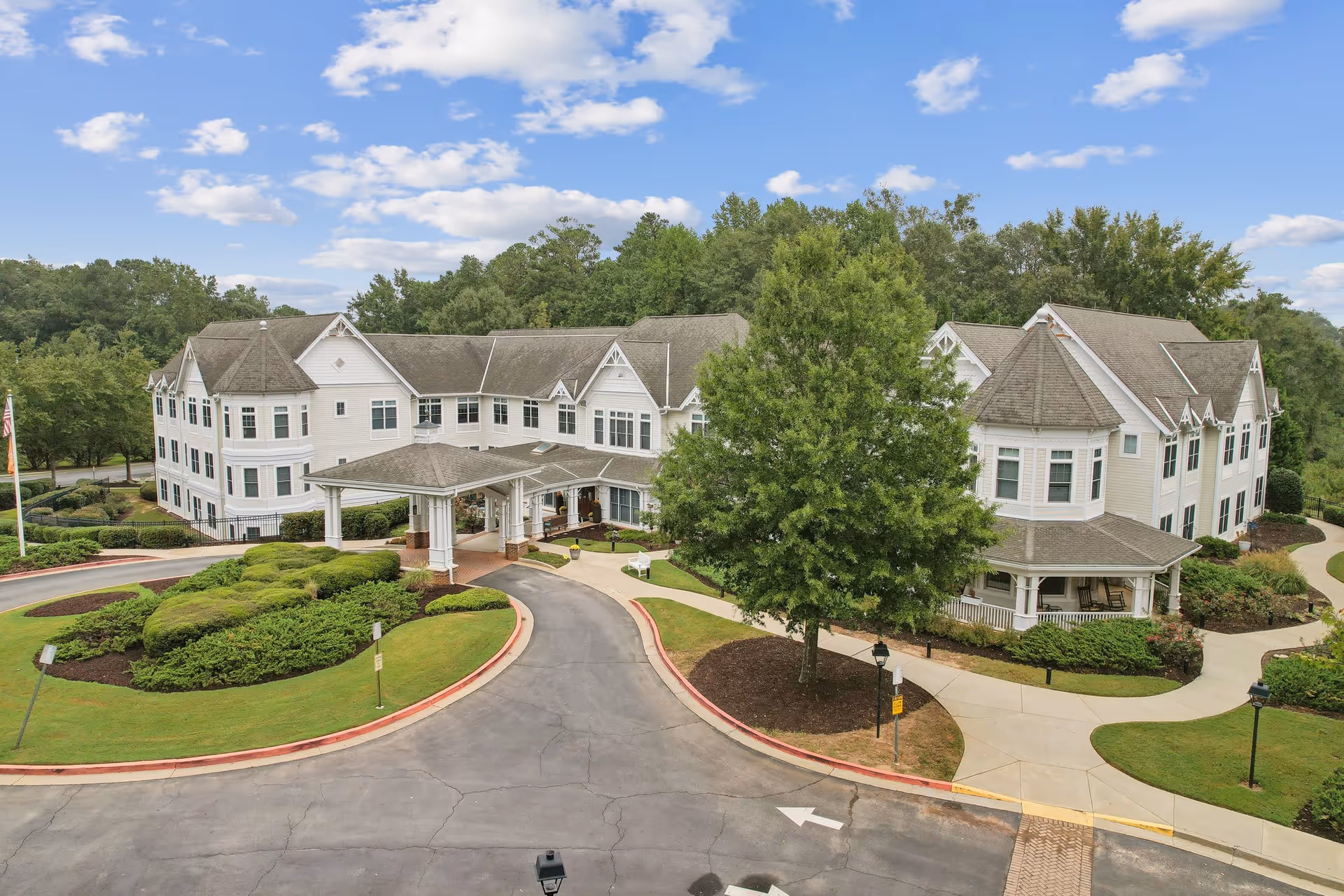 Exterior view of a large, white, multi-story assisted living facility building with a covered entrance driveway, surrounded by well-maintained landscaping and trees under a blue sky with scattered clouds.