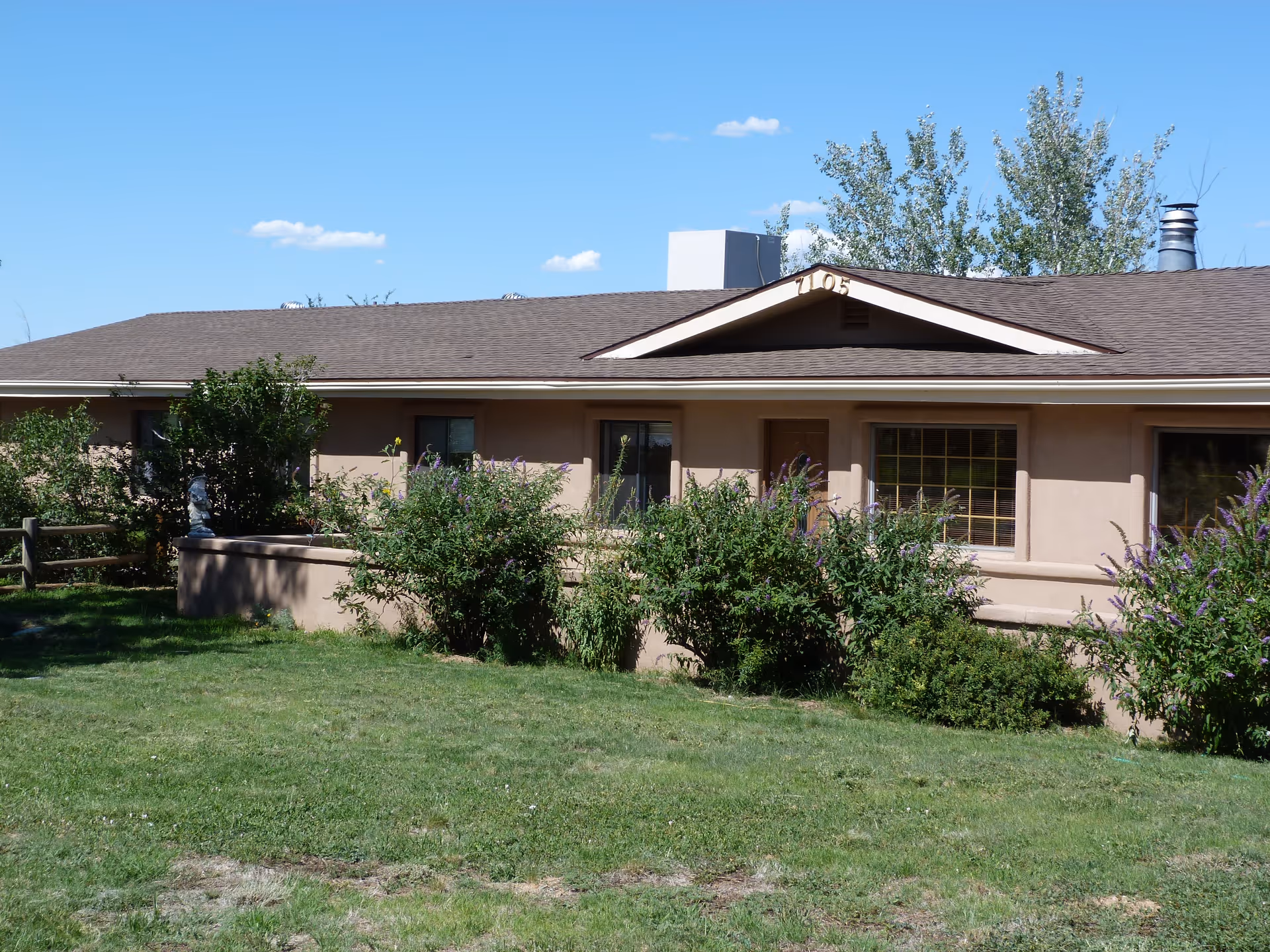 Single-story beige building with a brown shingled roof, surrounded by green bushes and grass under a clear blue sky. The building has several windows and a door, with the number 1105 displayed near the roof peak.