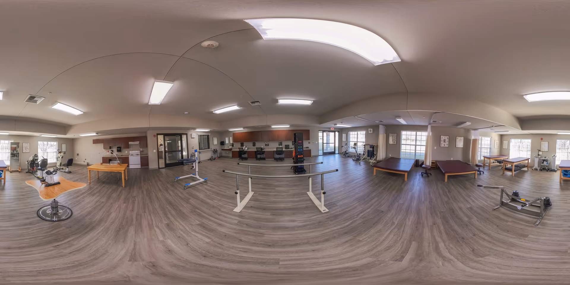 Wide-angle interior of a rehabilitation/exercise room with parallel bars, therapy tables, and fitness equipment.