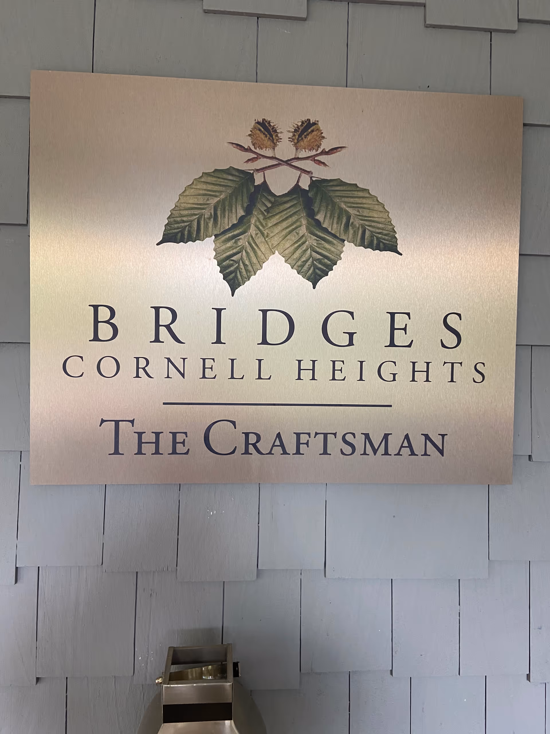Copper-toned sign reading Bridges Cornell Heights The Craftsman mounted on a gray shingle wall.