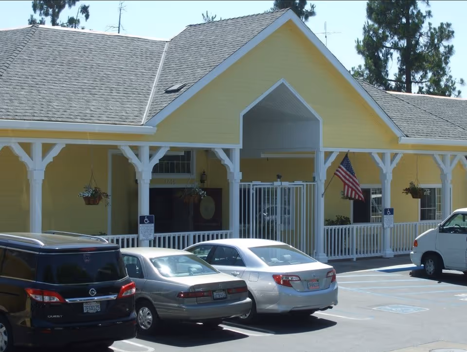 Yellow one-story building with a white porch, American flag, hanging plants, and multiple cars parked in front.