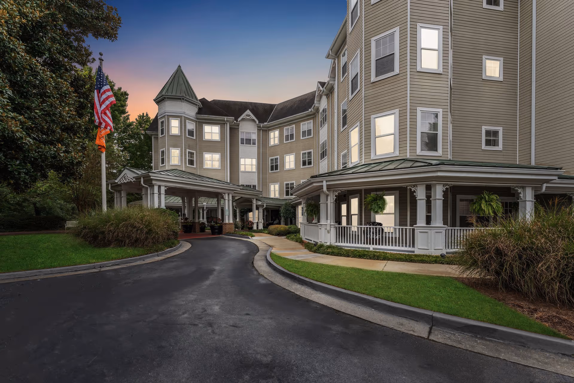 Exterior view of a multi-story senior living facility building at sunset with a covered entrance driveway, manicured lawns, and American and orange flags on a flagpole.