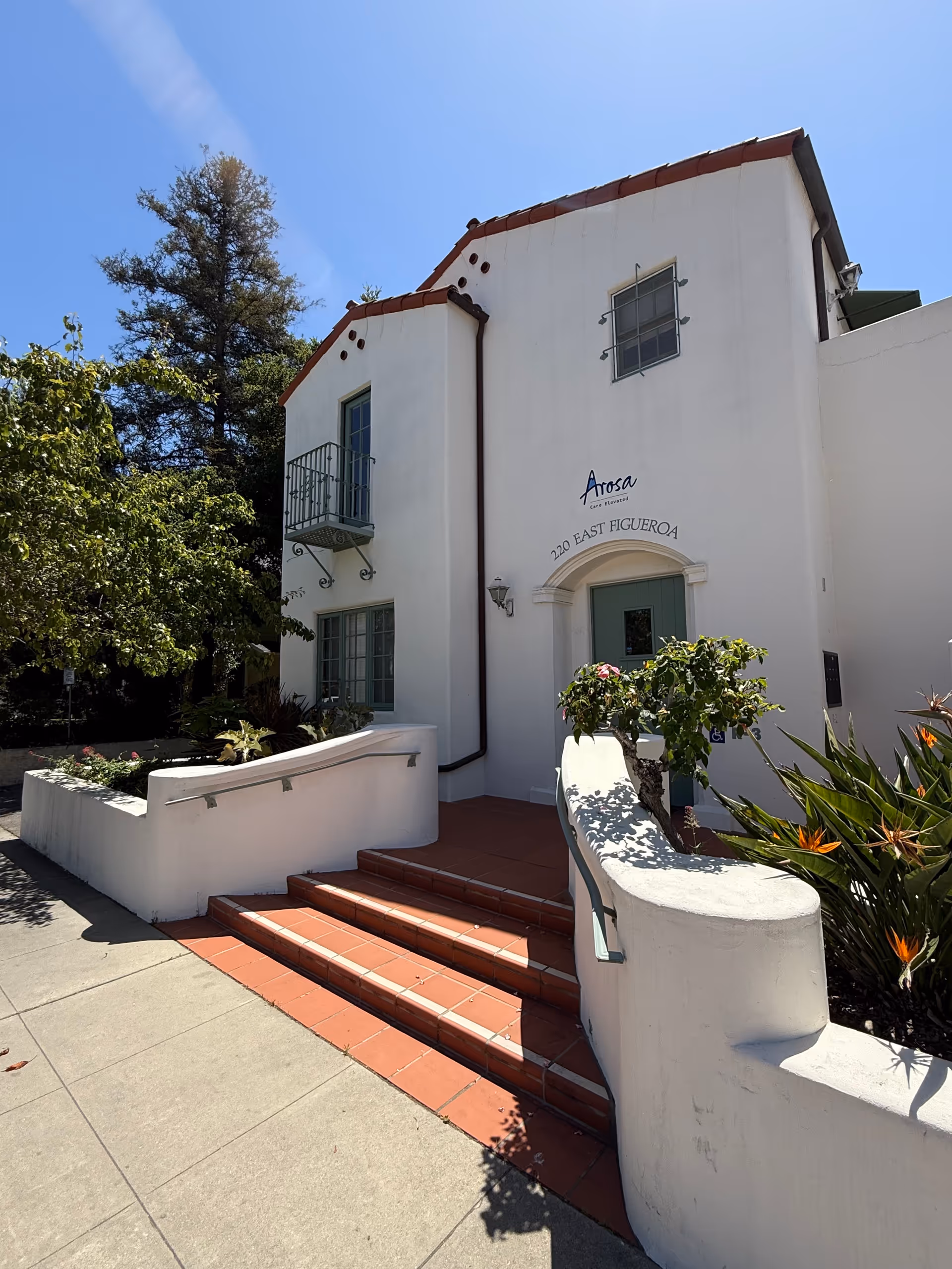 White stucco senior living building with red-tiled stairs, balcony, and surrounding greenery.