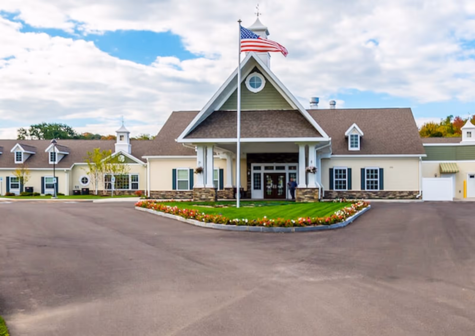 Front exterior of a single-story senior living facility with a central entrance canopy, flagpole, and landscaped driveway.