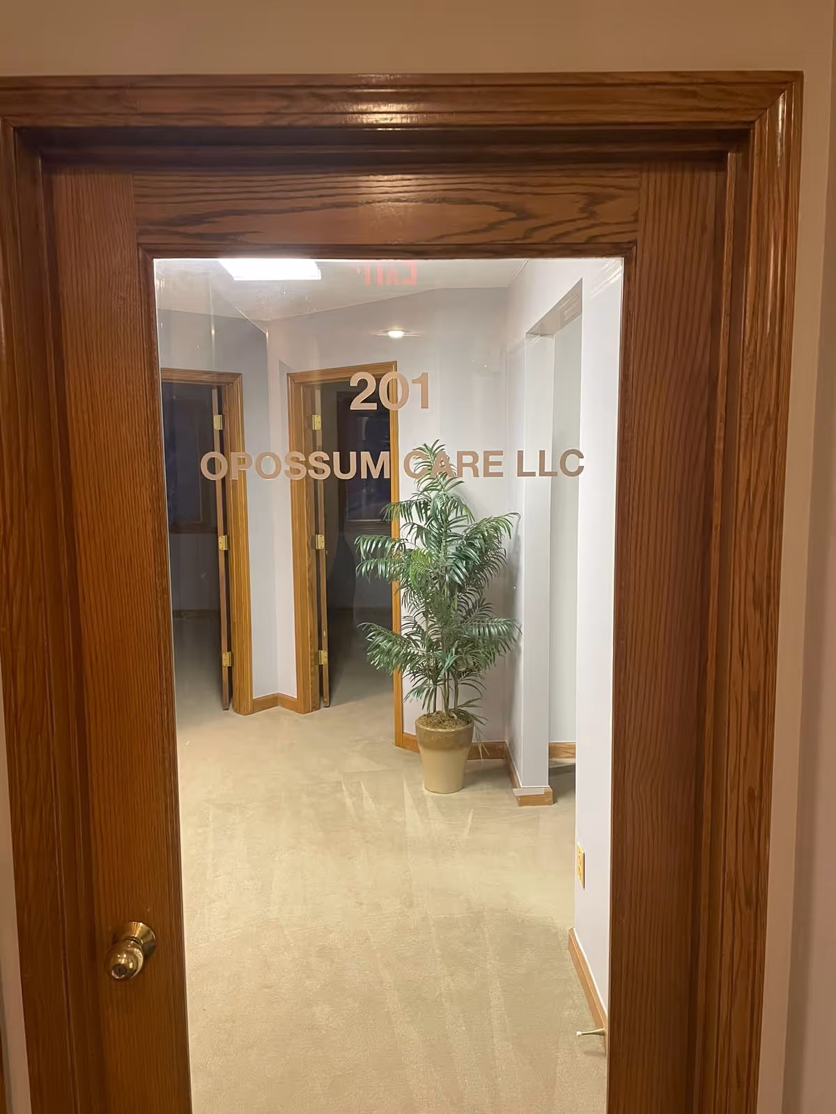 Wood-framed glass door bearing Opossum Care LLC signage and a potted plant in a hallway.