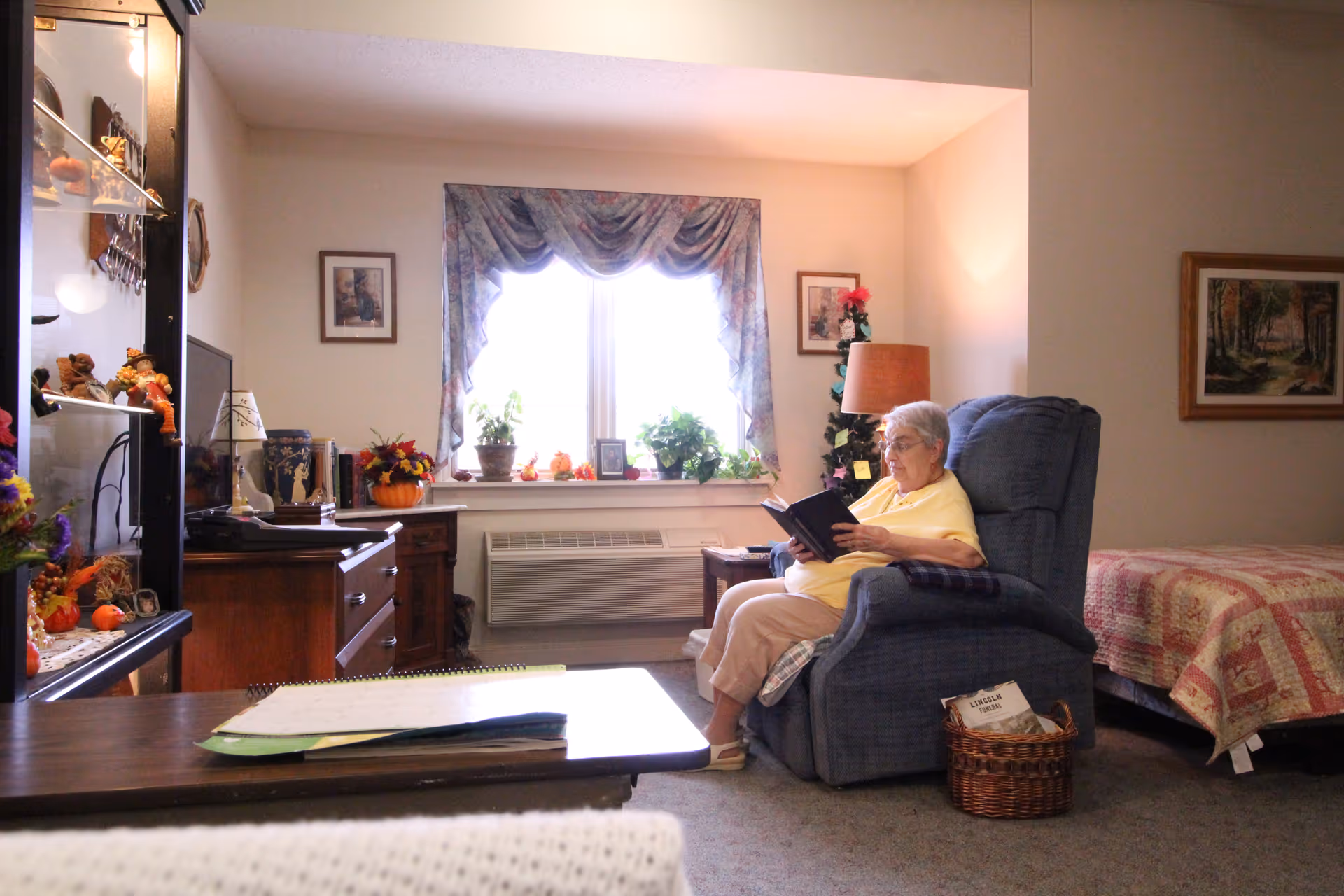 An elderly woman sitting in a blue recliner chair reading a book in a cozy room with a bed covered in a quilt, a window with floral curtains, and various decorations including plants, framed pictures, and a lamp.