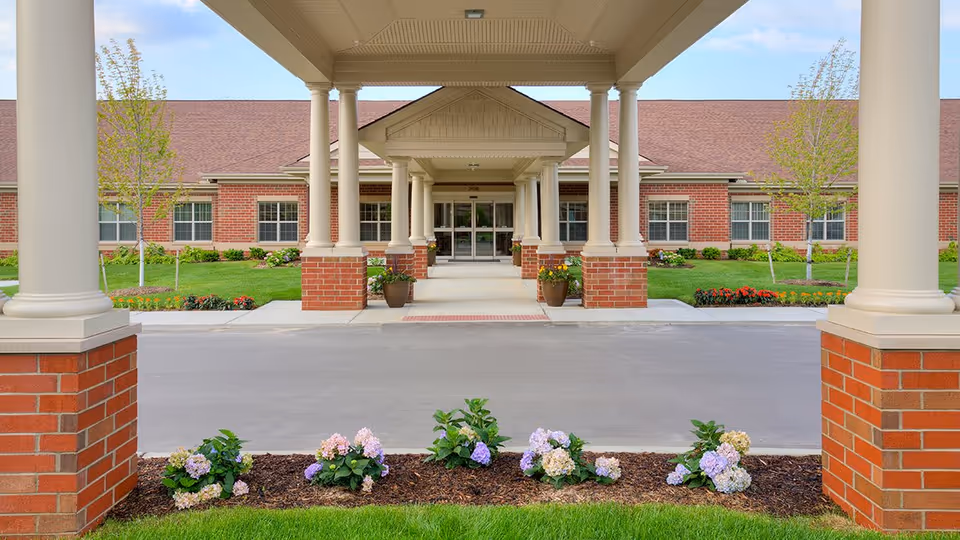 Front entrance of a brick assisted living building with a covered portico supported by columns and landscaped flower beds.