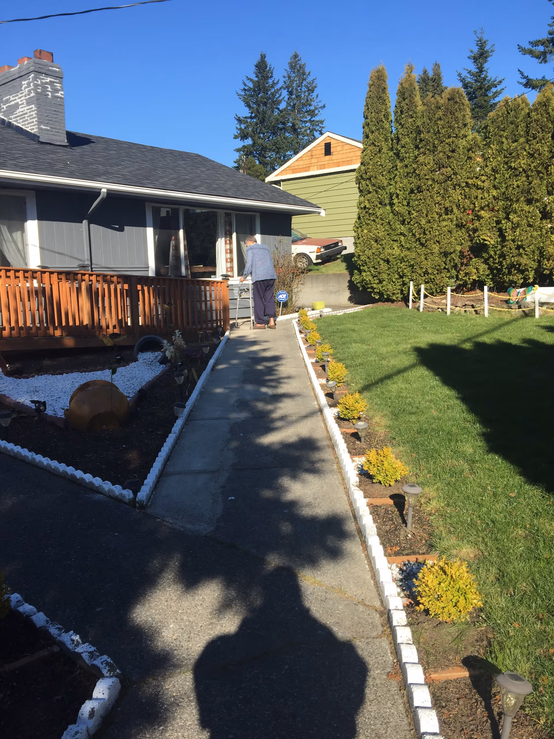 Elderly person standing on a walkway toward a single-story home with a wooden railing and shrubs.
