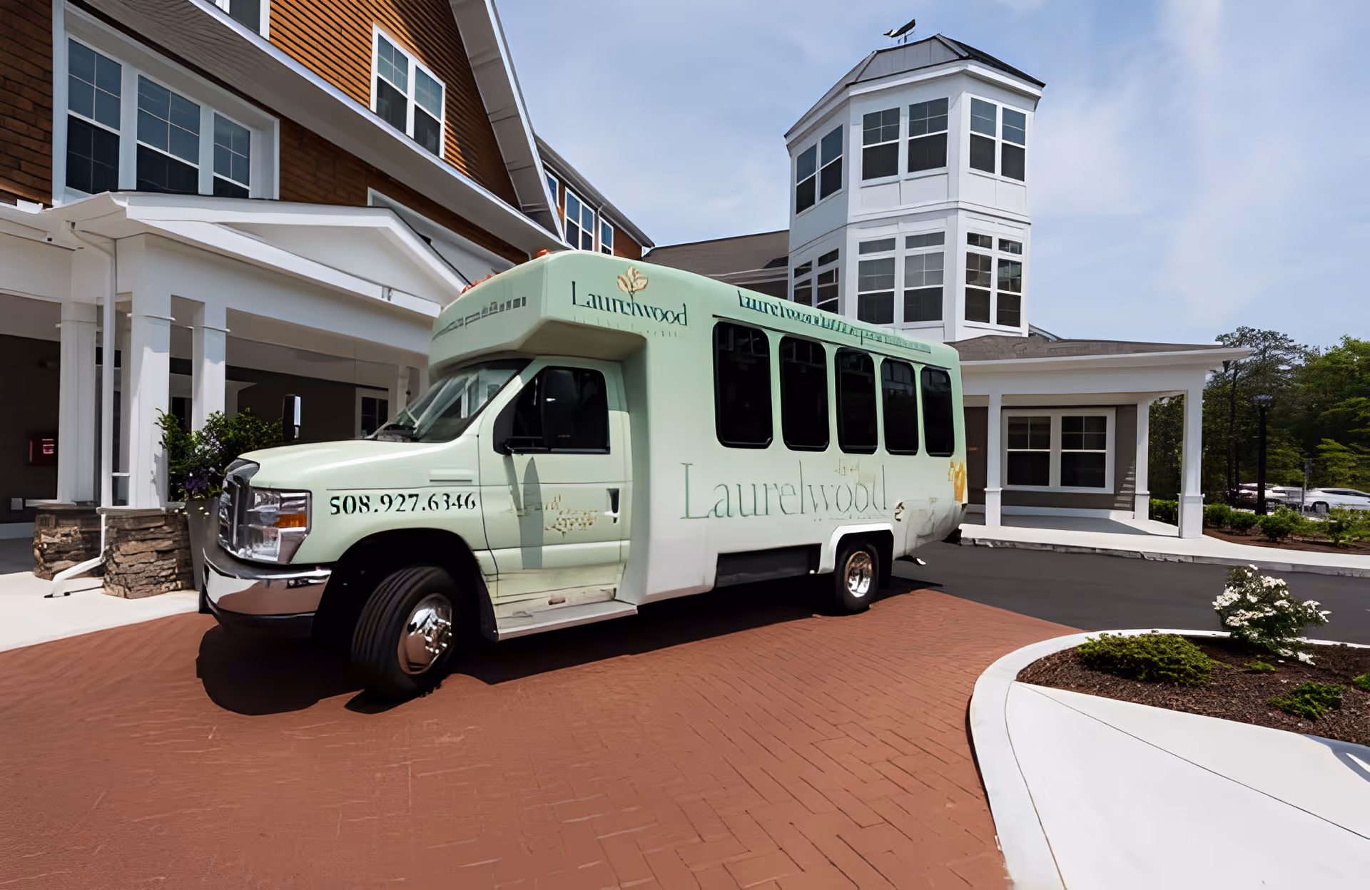 A light green shuttle bus with the name 'Laurelwood' parked in front of a senior living facility building with white columns and a multi-windowed tower section under a clear blue sky.