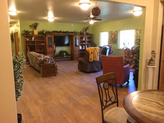 Spacious assisted-living common room with sofas and armchairs facing a TV entertainment center, plants, and wood-look flooring.
