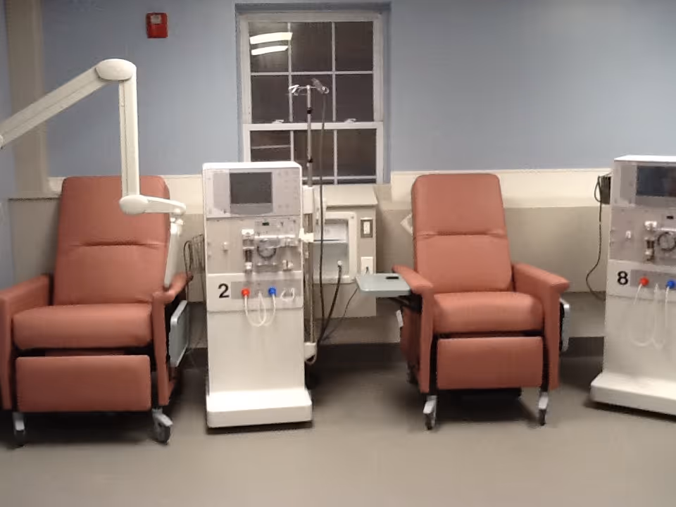 A medical treatment room with two brown recliner chairs positioned next to dialysis machines. The room has a window in the background and light blue walls with a white chair rail.