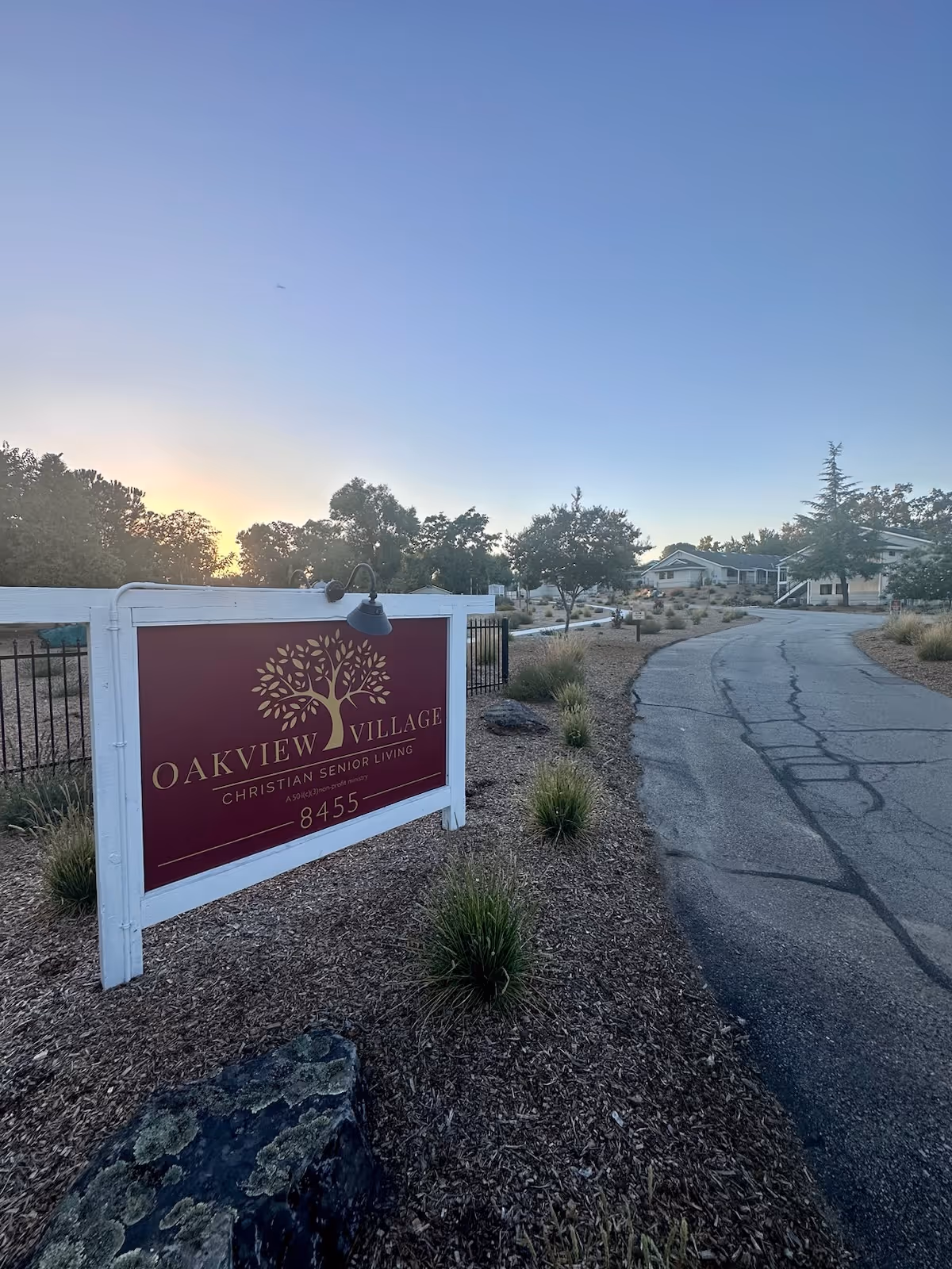 Outdoor view of Oakview Village Independent Senior Living facility entrance sign with a paved driveway leading to buildings surrounded by trees and landscaping under a clear sky at sunset.