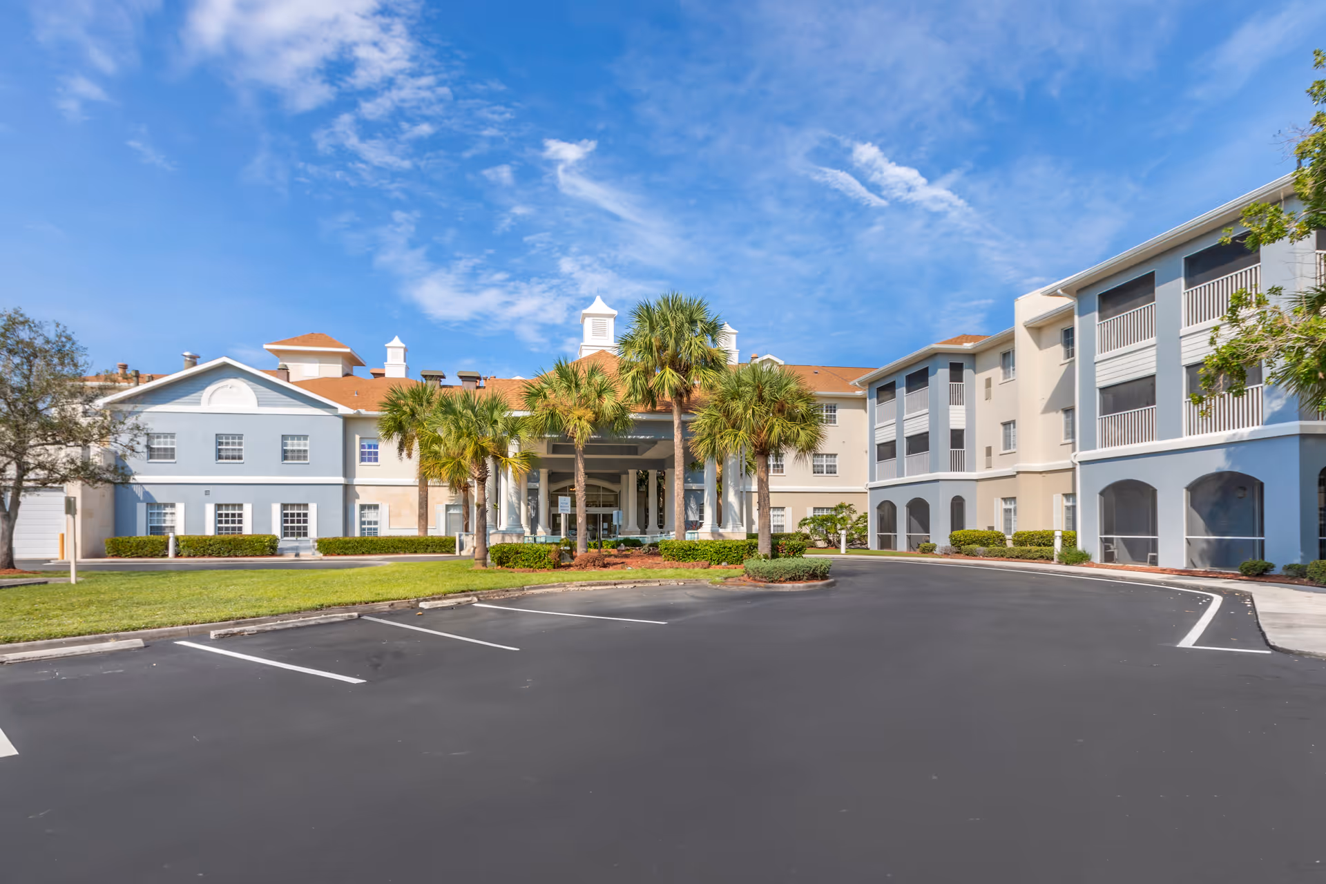 Exterior view of Brookdale Fort Myers Cypress Lake senior living facility showing a large building with multiple floors, palm trees in front, a covered entrance with white columns, and a clear blue sky above.