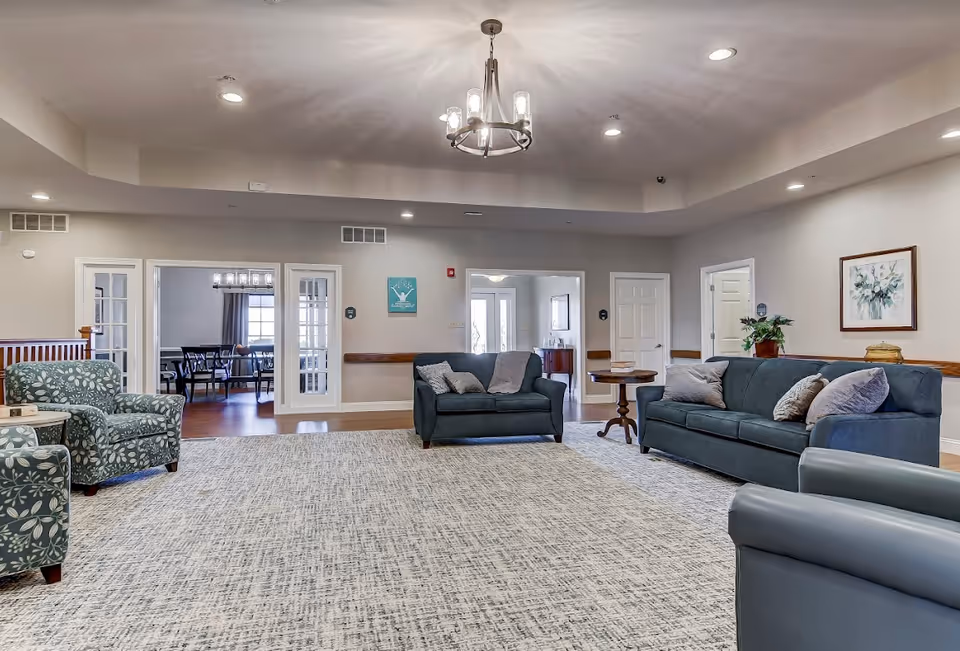 Spacious senior living common room with sofas, armchairs, a large rug, and a dining area visible through glass doors.