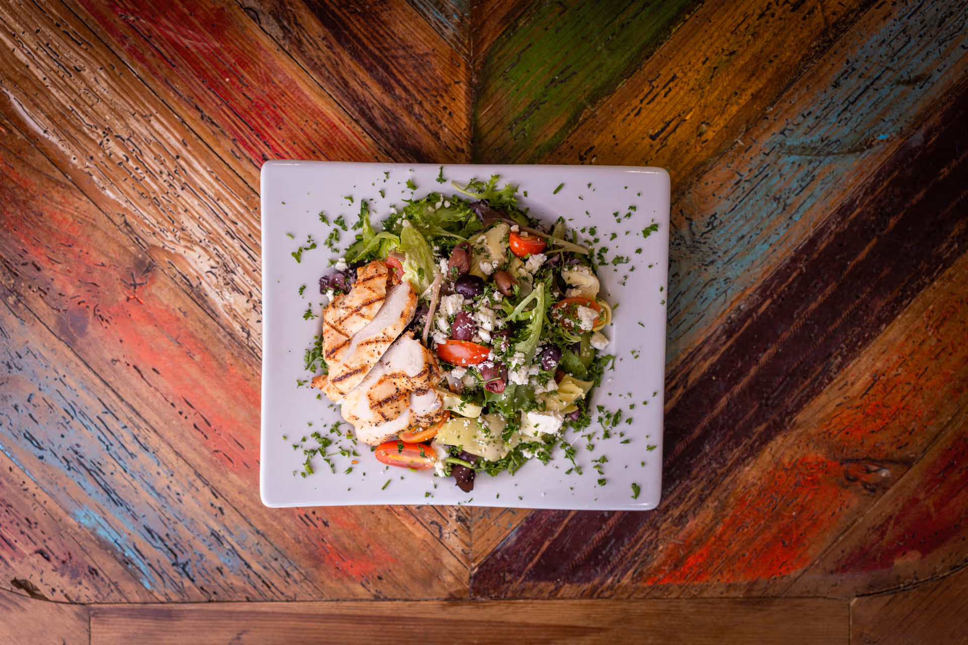A white square plate with a grilled chicken breast and a mixed salad containing lettuce, cherry tomatoes, olives, artichokes, and crumbled cheese, garnished with chopped herbs, placed on a colorful wooden table with a chevron pattern.