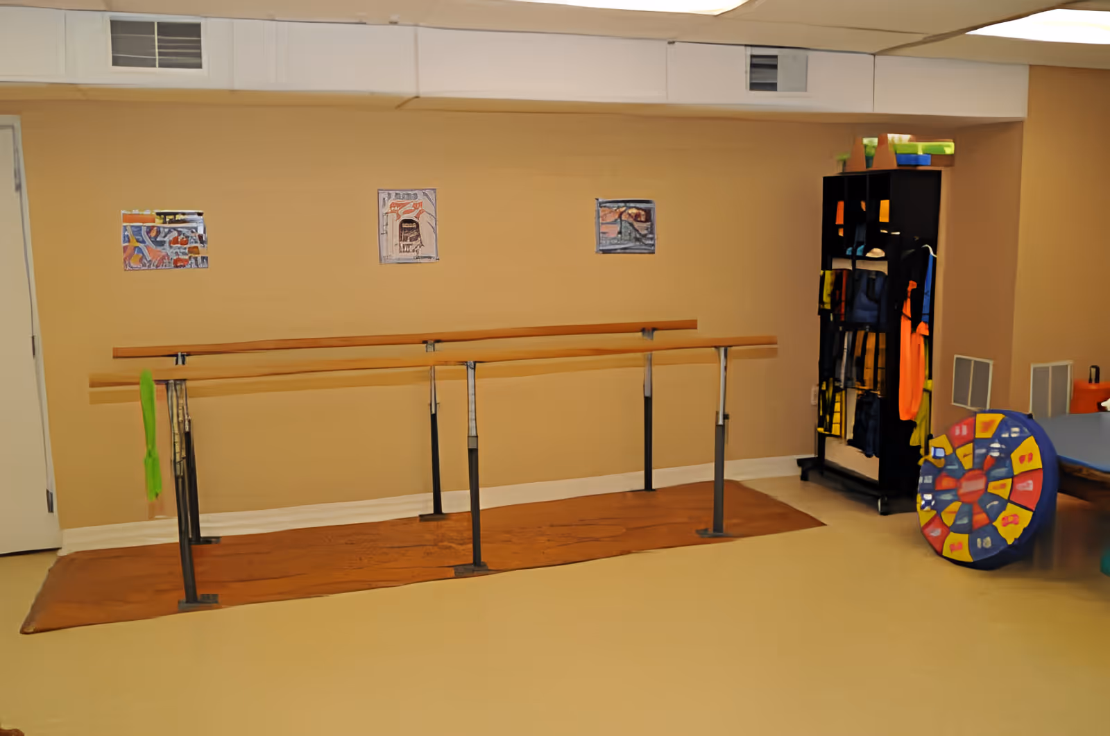 A therapy or exercise room with parallel wooden handrails mounted on metal supports along a beige wall. There are three small colorful artworks hanging on the wall above the handrails. To the right, there is a black shelving unit with various colorful items and a large circular colorful padded object leaning against it. The floor is light-colored and there is a brown mat under the handrails.