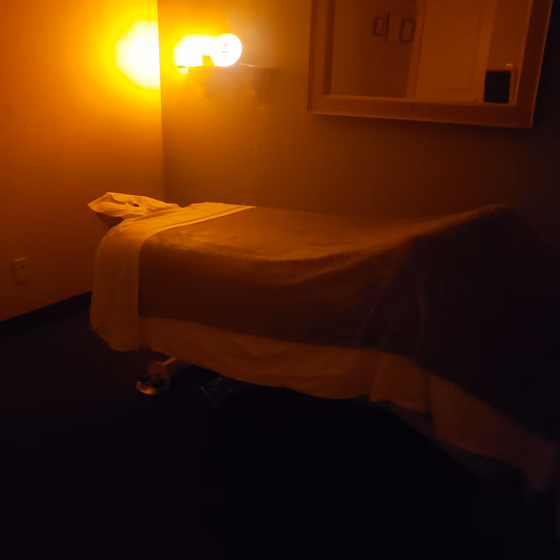 Dimly lit bedroom with a single bed, warm yellow wall lamp, and mirror on the wall.
