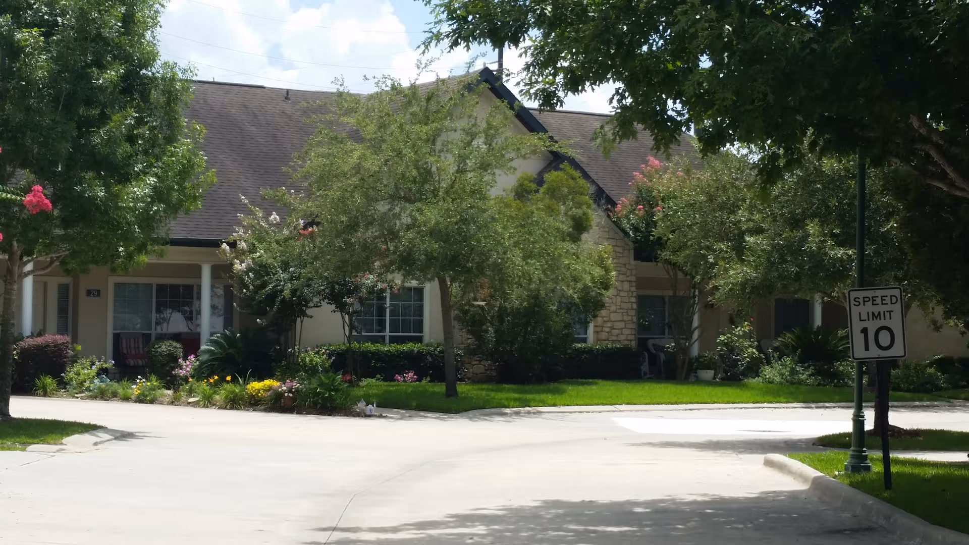 Exterior view of a senior living facility with front lawn, trees, and a speed limit sign.