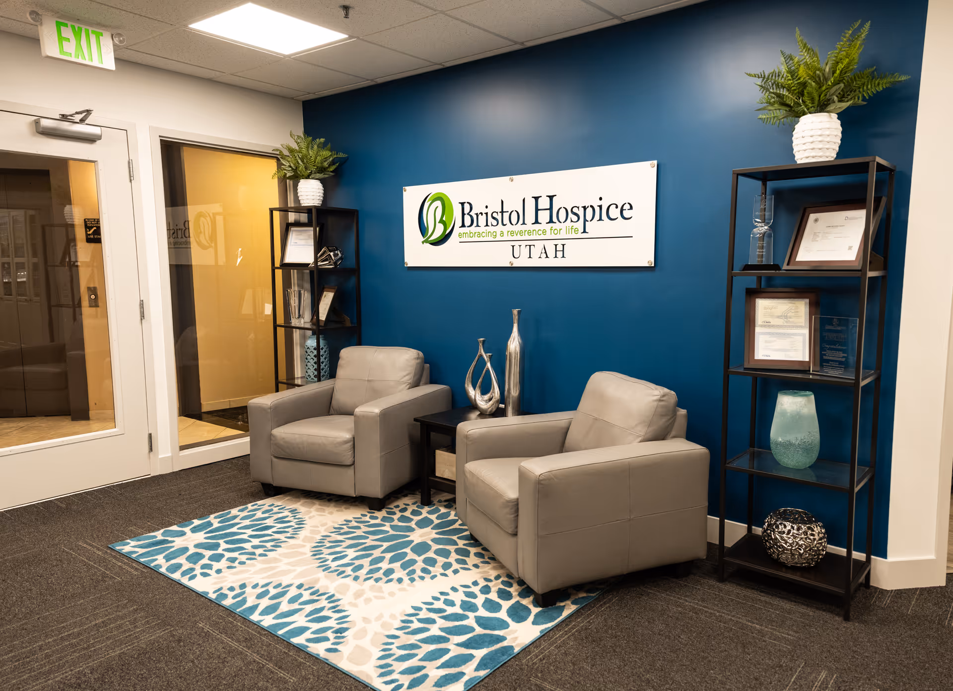 Reception area with two gray chairs, teal blue wall, Bristol Hospice Utah sign, and shelves.