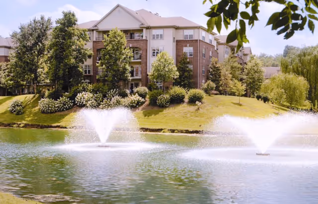 Brick senior living building by a landscaped pond with fountains and trees.