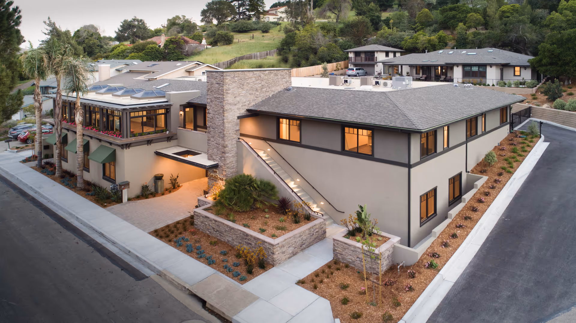 A modern two-story elder care facility with stone accents, landscaped courtyards, and a paved road.