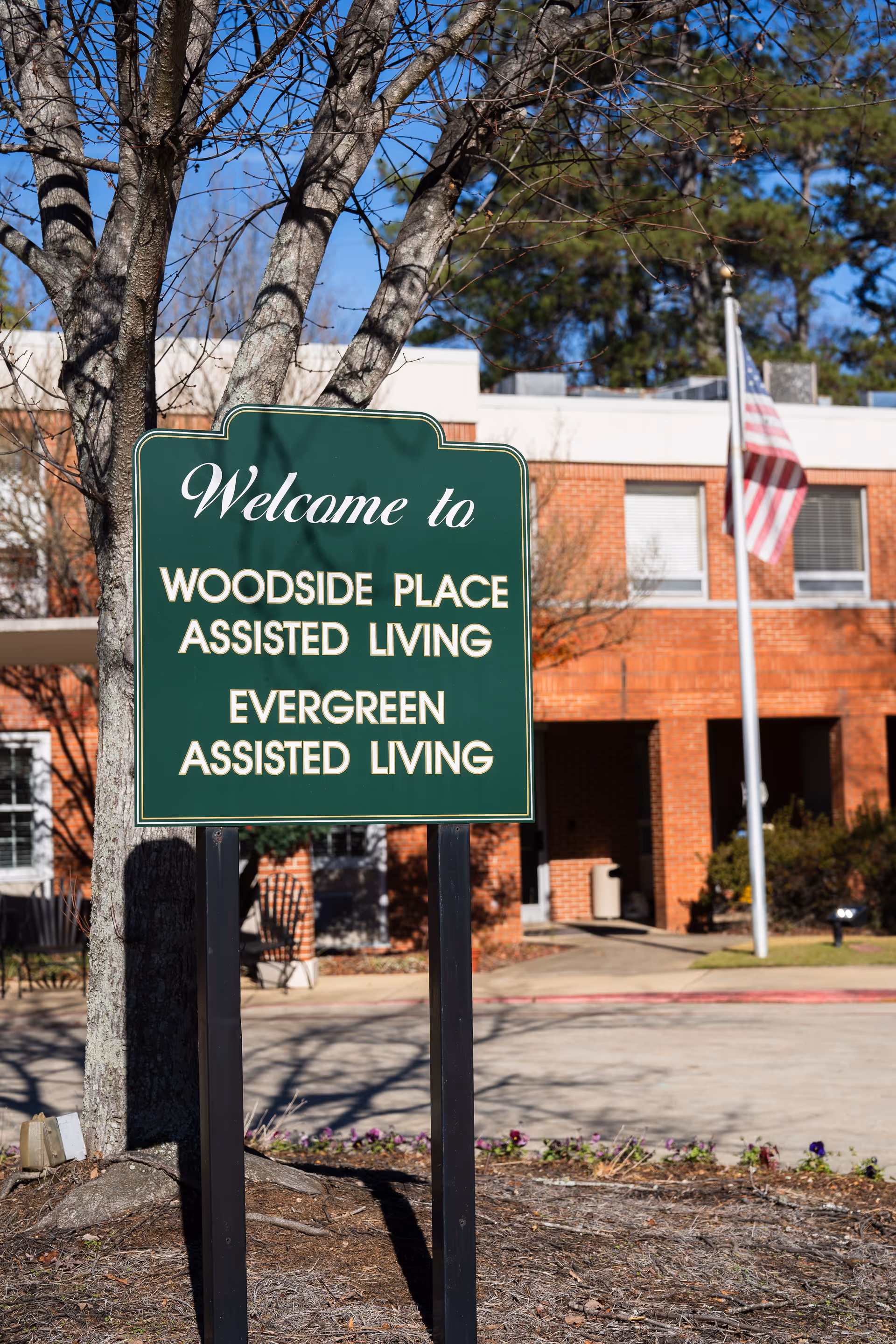 Green sign reads Woodside Place Assisted Living in front of a brick building.