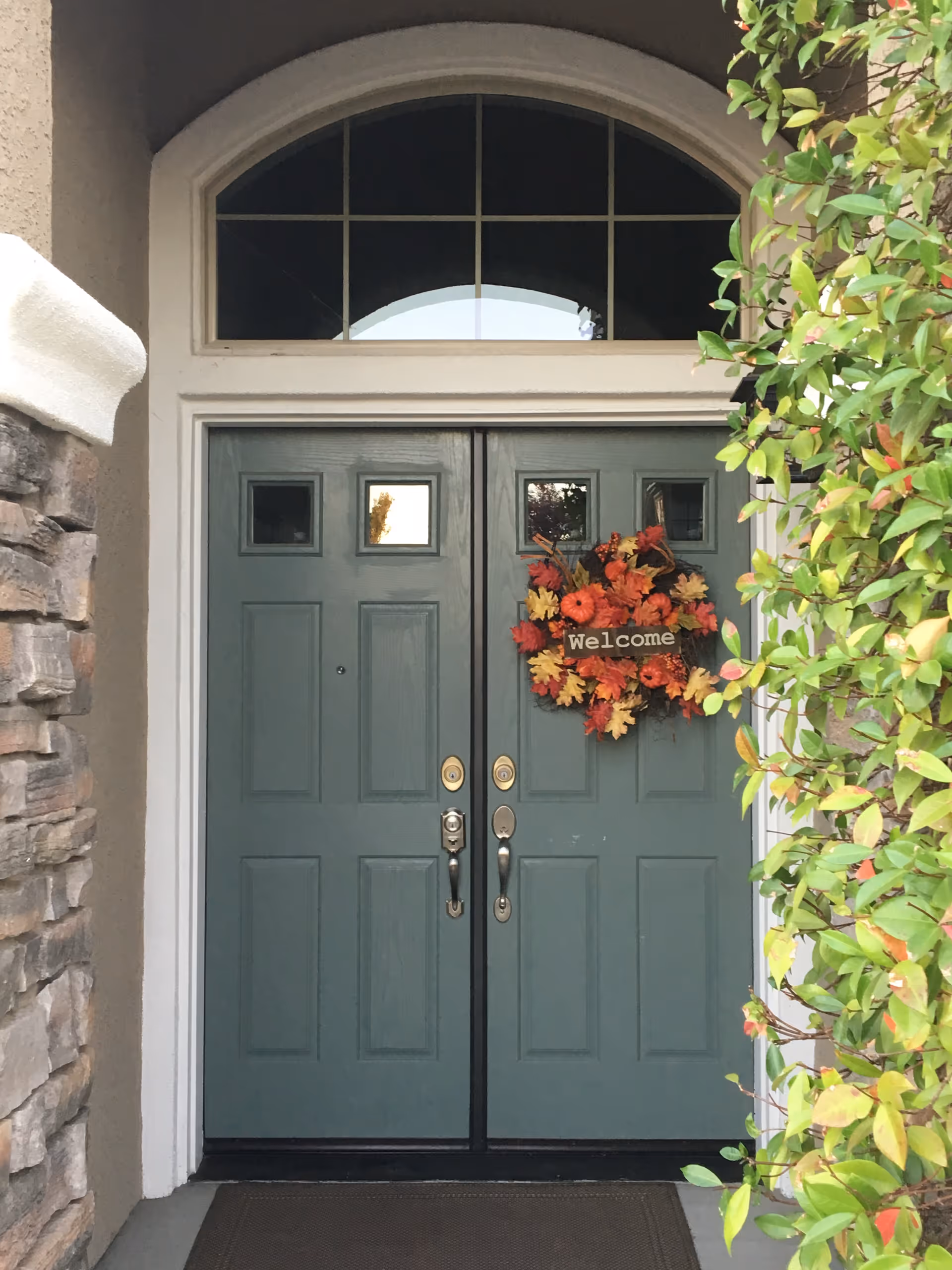 Double teal front doors with a fall wreath reading 'Welcome', flanked by a stone wall on the left and leafy greenery on the right.