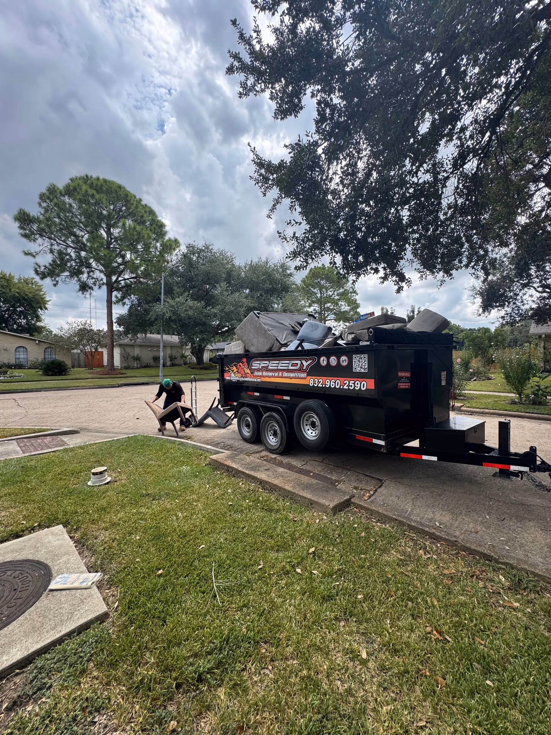 Black dumpster-style trailer overflowing with furniture on a residential street; a person stacks items nearby.