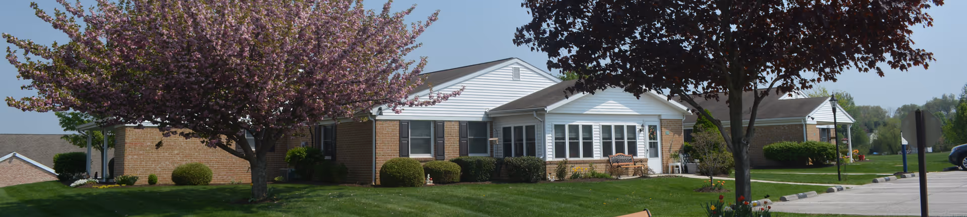 Exterior view of a single-story brick and white siding building with multiple windows, surrounded by green grass, bushes, and blooming trees with pink and dark red leaves under a clear blue sky.