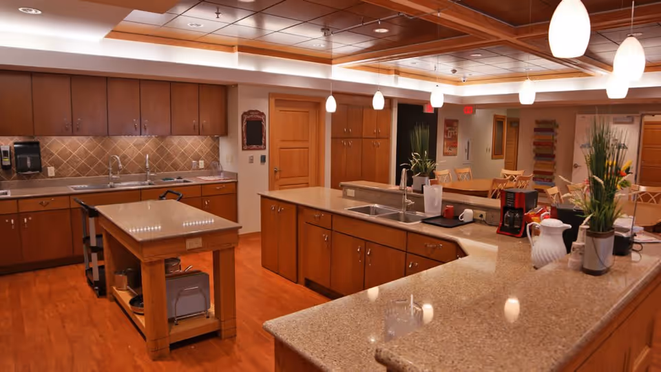 A spacious, well-lit kitchen area with wooden cabinets, granite countertops, and multiple sinks. There is a central island with storage underneath, pendant lights hanging from the ceiling, and various kitchen items including a coffee maker and plants on the counters. The floor is wooden, and there are doors and additional cabinetry in the background.
