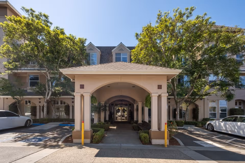 Entrance porte-cochere of a multi-story senior living community building flanked by trees and parked cars.