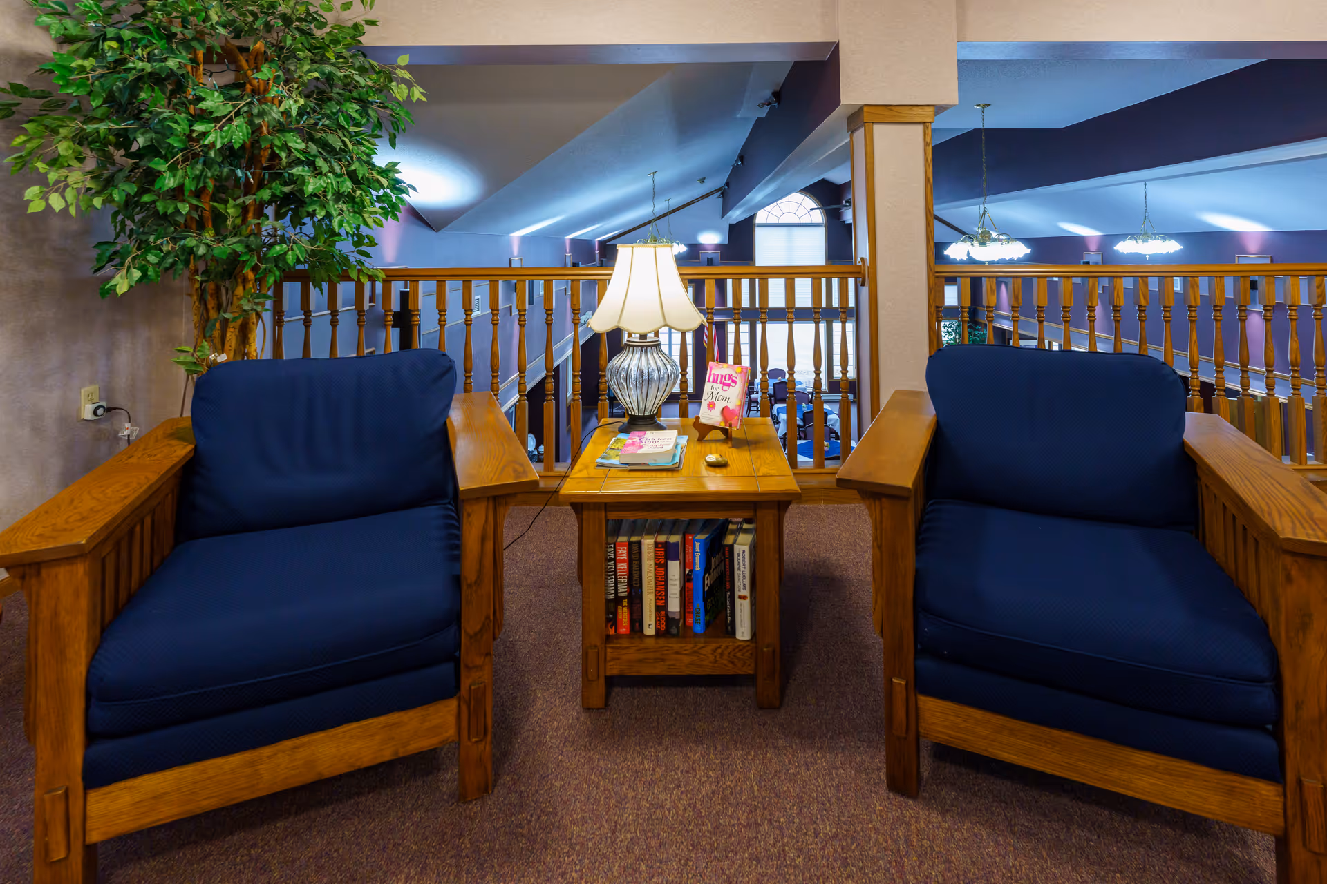 Two wooden armchairs with dark blue cushions flank a wooden side table with a lamp, books, and a card on it. Behind the seating area is a wooden railing overlooking a lower level with hanging lights and a large window.