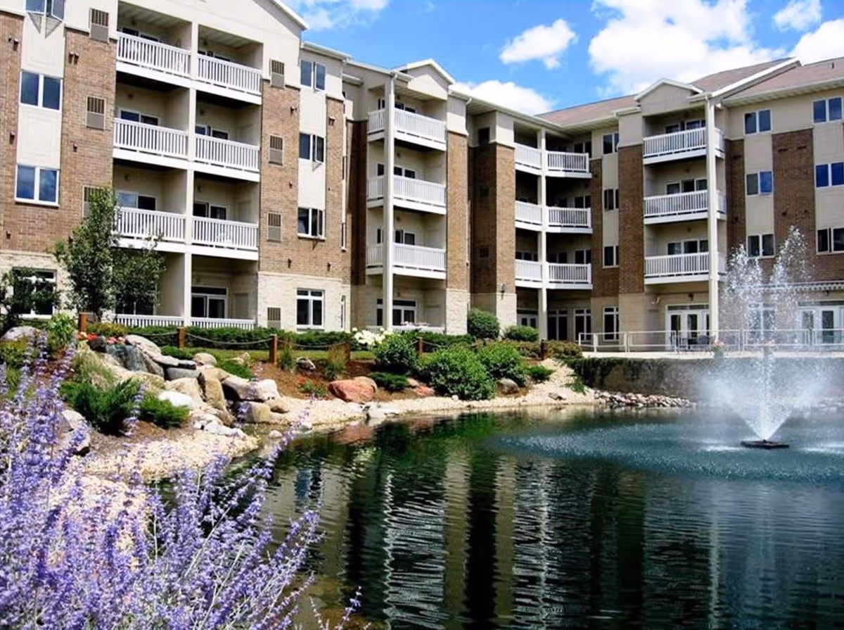 Exterior view of a multi-story senior living facility named Coventry Village with balconies overlooking a landscaped pond featuring a water fountain and surrounded by greenery and purple flowers under a partly cloudy sky.