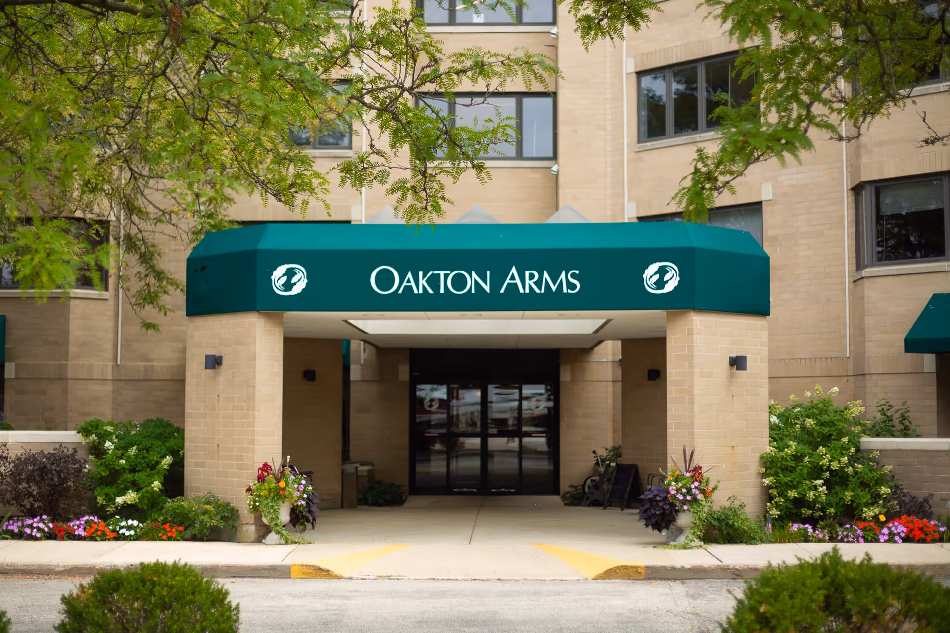 Entrance to a building with a green awning that reads 'OAKTON ARMS', surrounded by plants and flowers with beige brick walls and windows in the background.