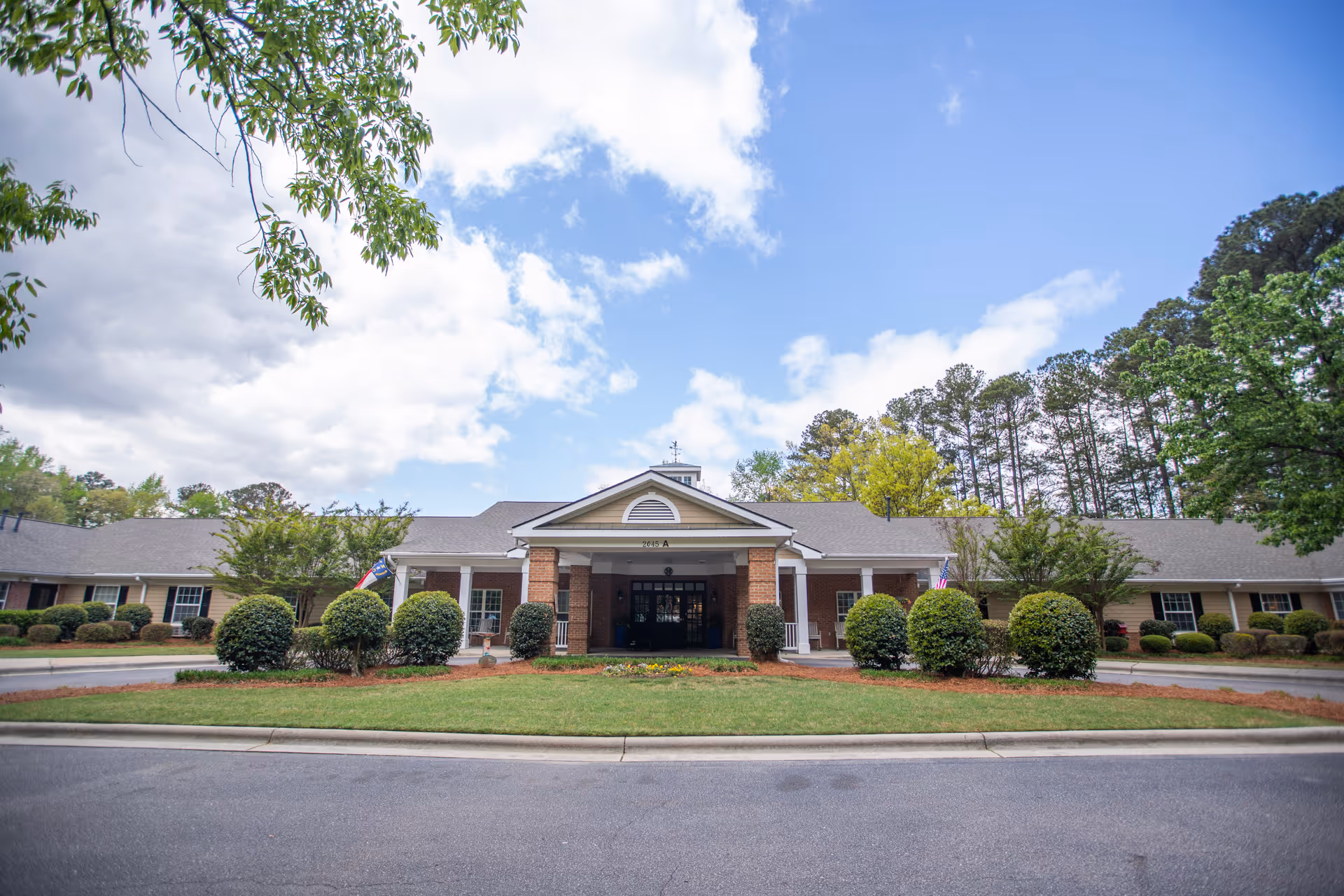 Front exterior view of a single-story senior living facility building with a covered entrance, surrounded by neatly trimmed bushes and trees under a partly cloudy blue sky.