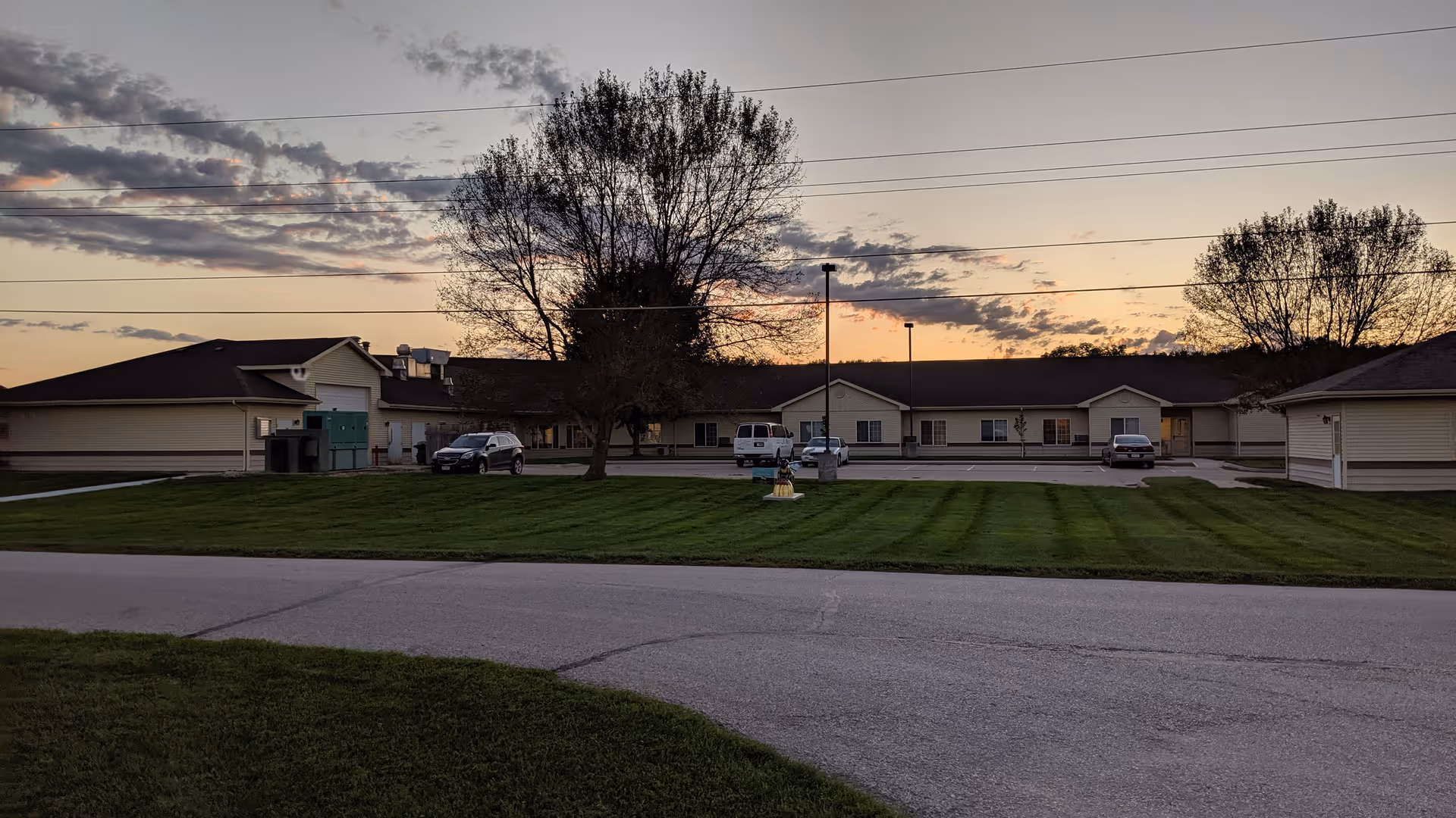Sunset exterior view of a single-story senior living campus with green lawn and parked cars.