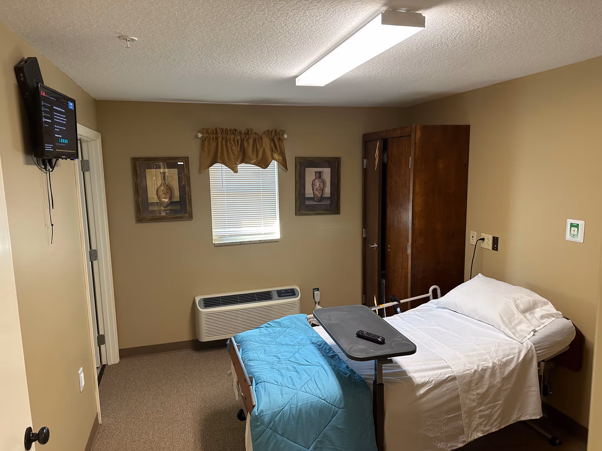 A small hospital-style bedroom with a bed, tray table, cabinet, and window blinds.
