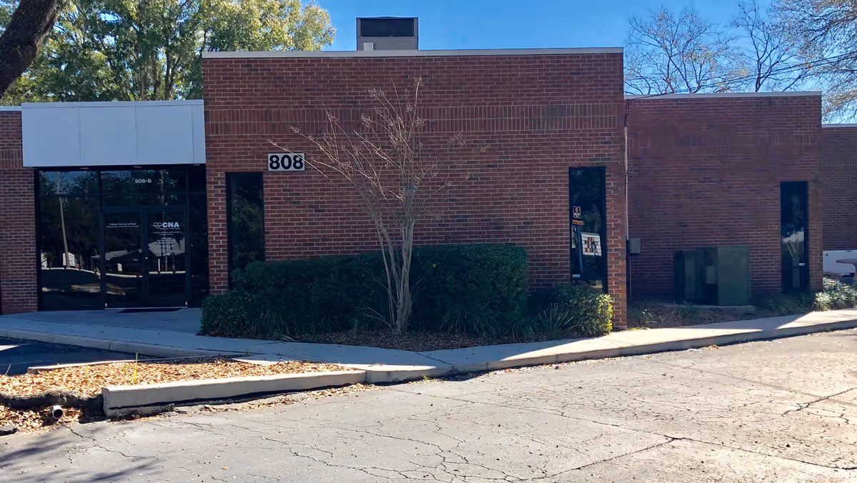 Brick building exterior with glass entry doors, sidewalk, and trimmed shrubs.