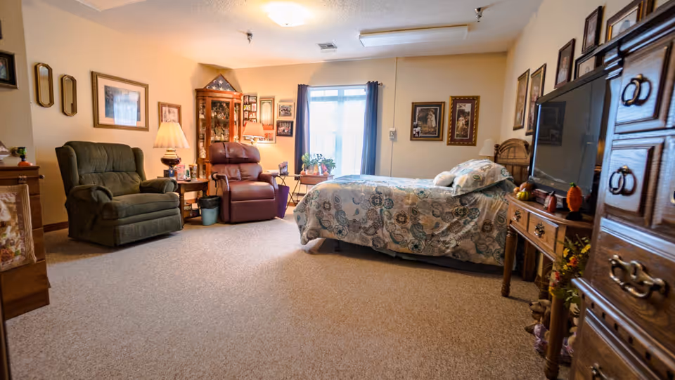 Carpeted senior living bedroom with a bed, two recliner chairs, a TV on a dresser, and framed pictures on the walls.
