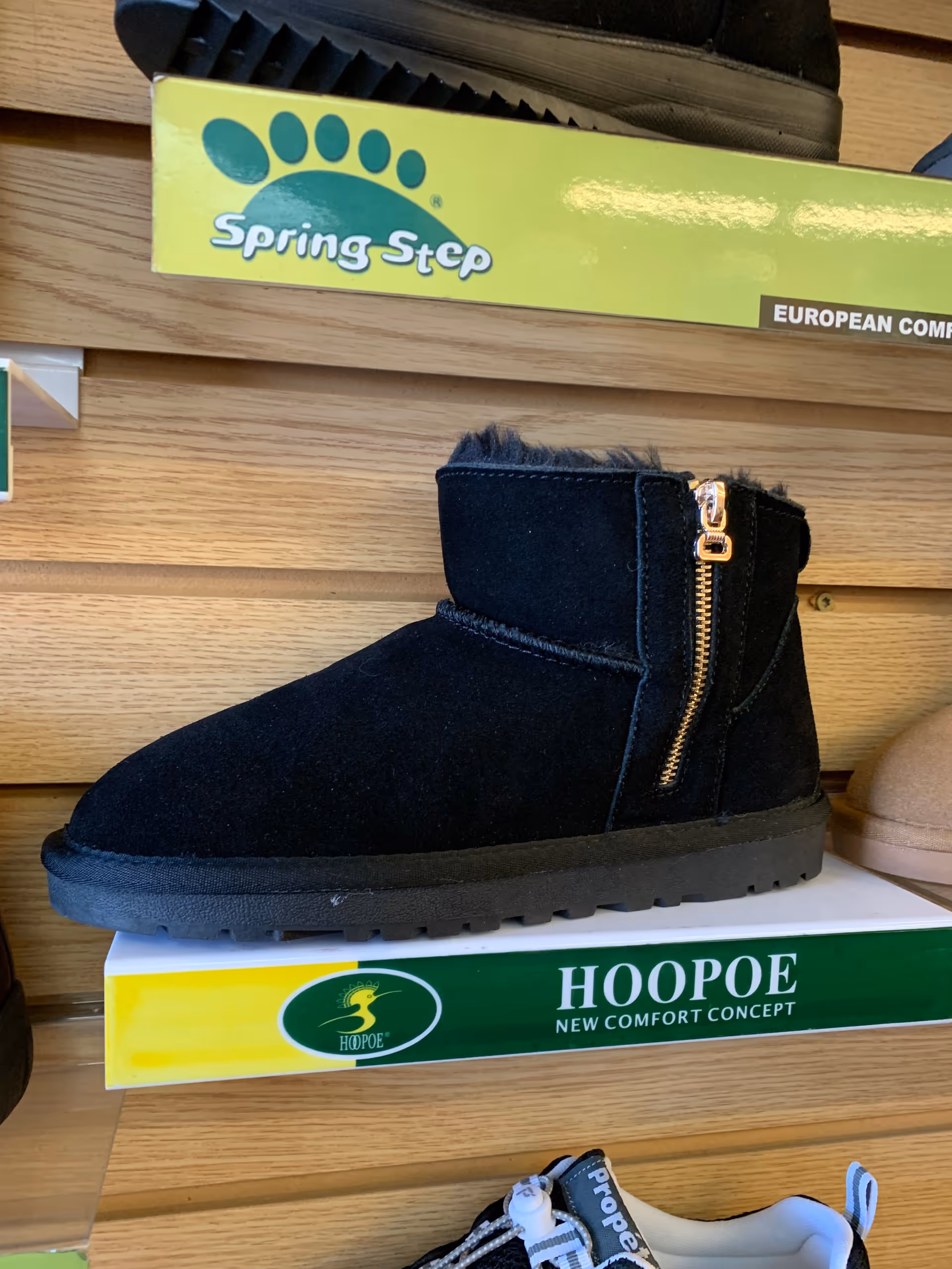 Black ankle boot with zipper displayed on a shelf among shoe boxes in a retail display.