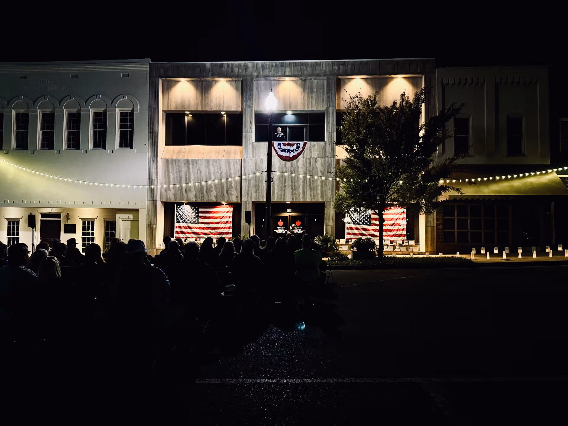 Night view of a storefront-like building with string lights, patriotic banners, and a seated crowd outside.