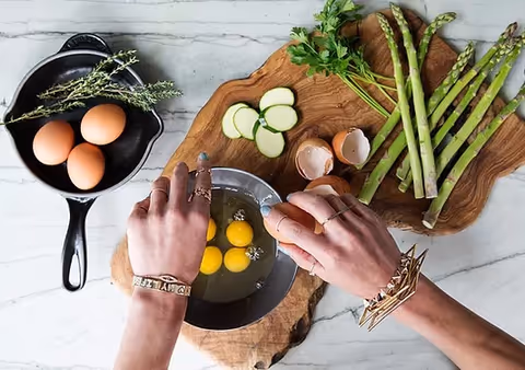 Two hands crack eggs into a bowl on a wooden board beside vegetables.