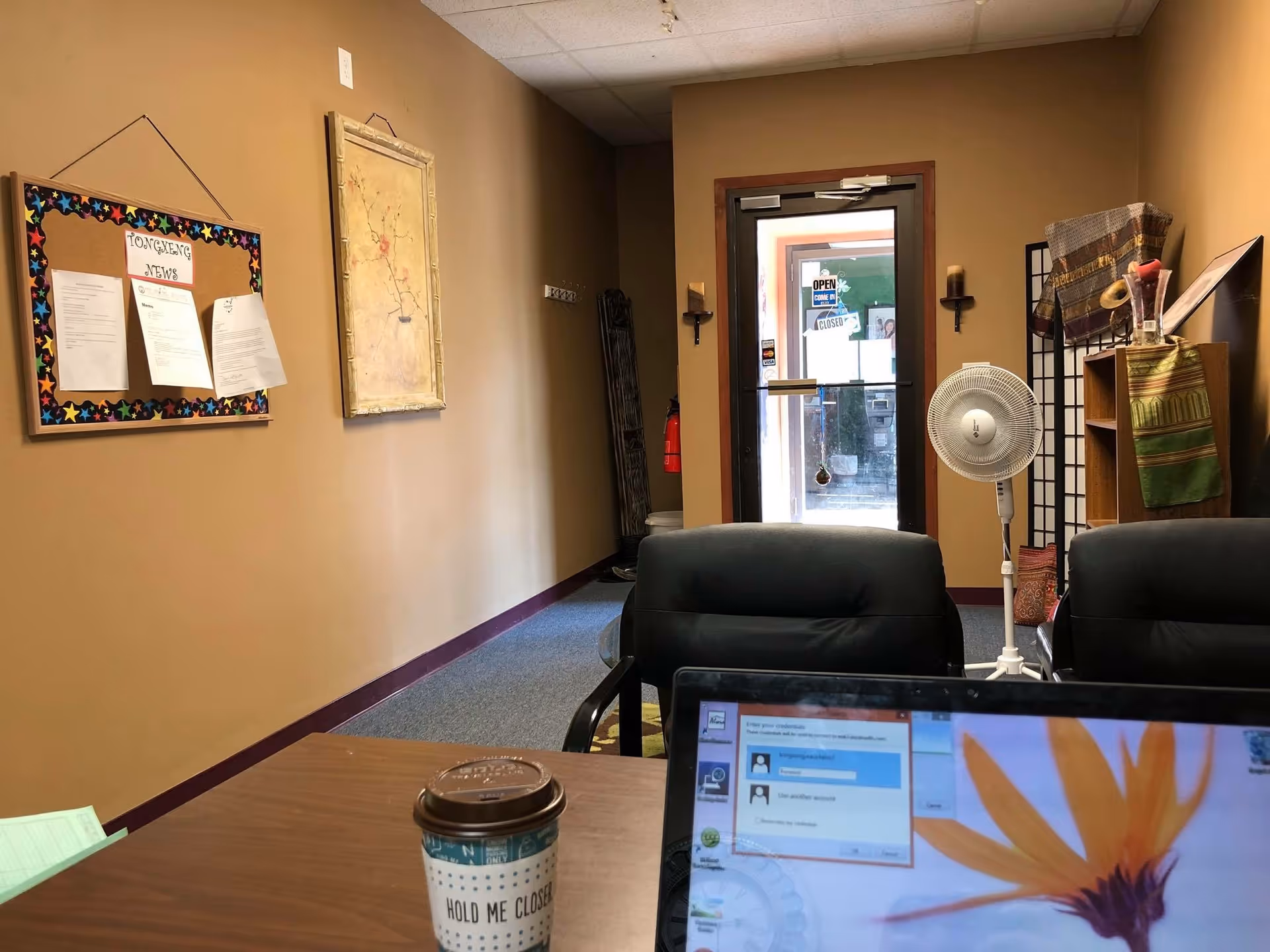 Office-like lobby with desk, laptop, coffee cup, fan, bulletin board, and doorway.