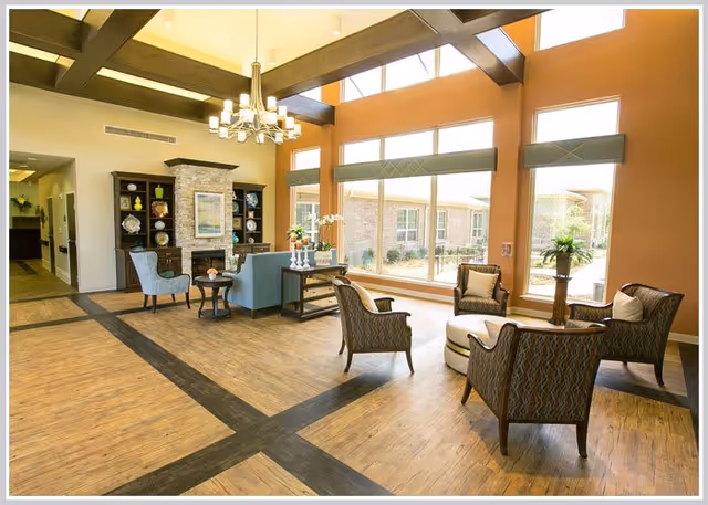 A bright and spacious living room area with large windows allowing natural light to fill the space. The room features a stone fireplace with a wooden mantel, a chandelier hanging from a high ceiling with exposed beams, and comfortable seating including armchairs and a sofa arranged around a round ottoman. Decorative shelves flank the fireplace, and there are plants and flowers adding a touch of greenery.